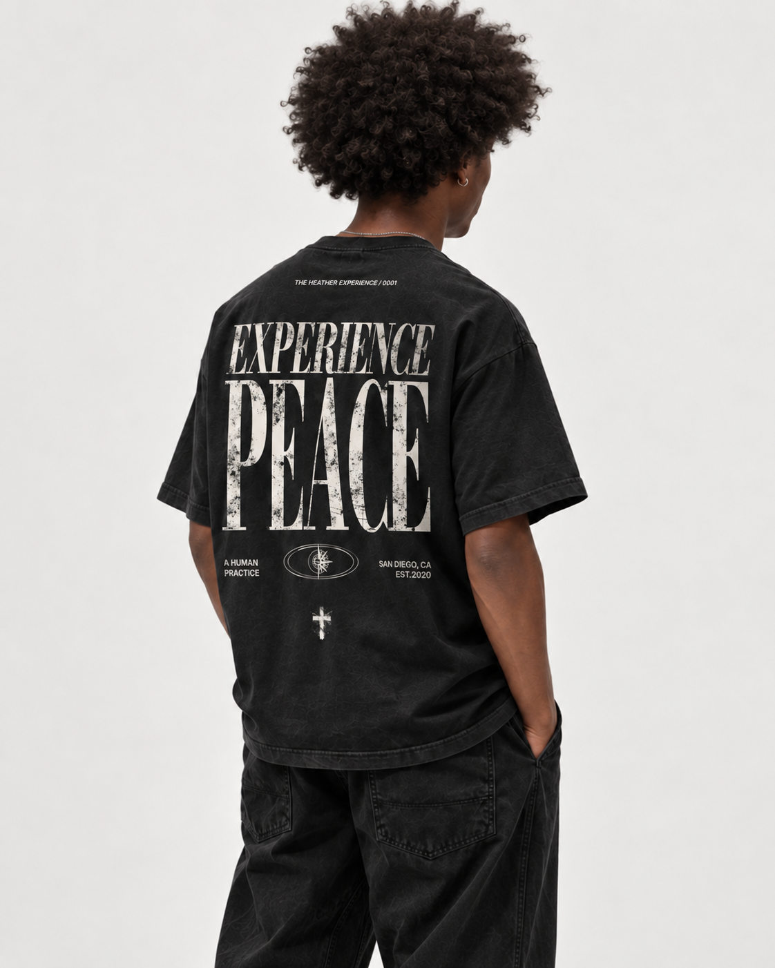 Back of a black EXPERIENCE PEACE T-shirt worn by a model, oversized distressed serif type reading THE HEATHER EXPERIENCE, EXPERIENCE PEACE, A HUMAN PRACTICE, an eye/compass emblem, and SAN DIEGO, CA EST. 2020.