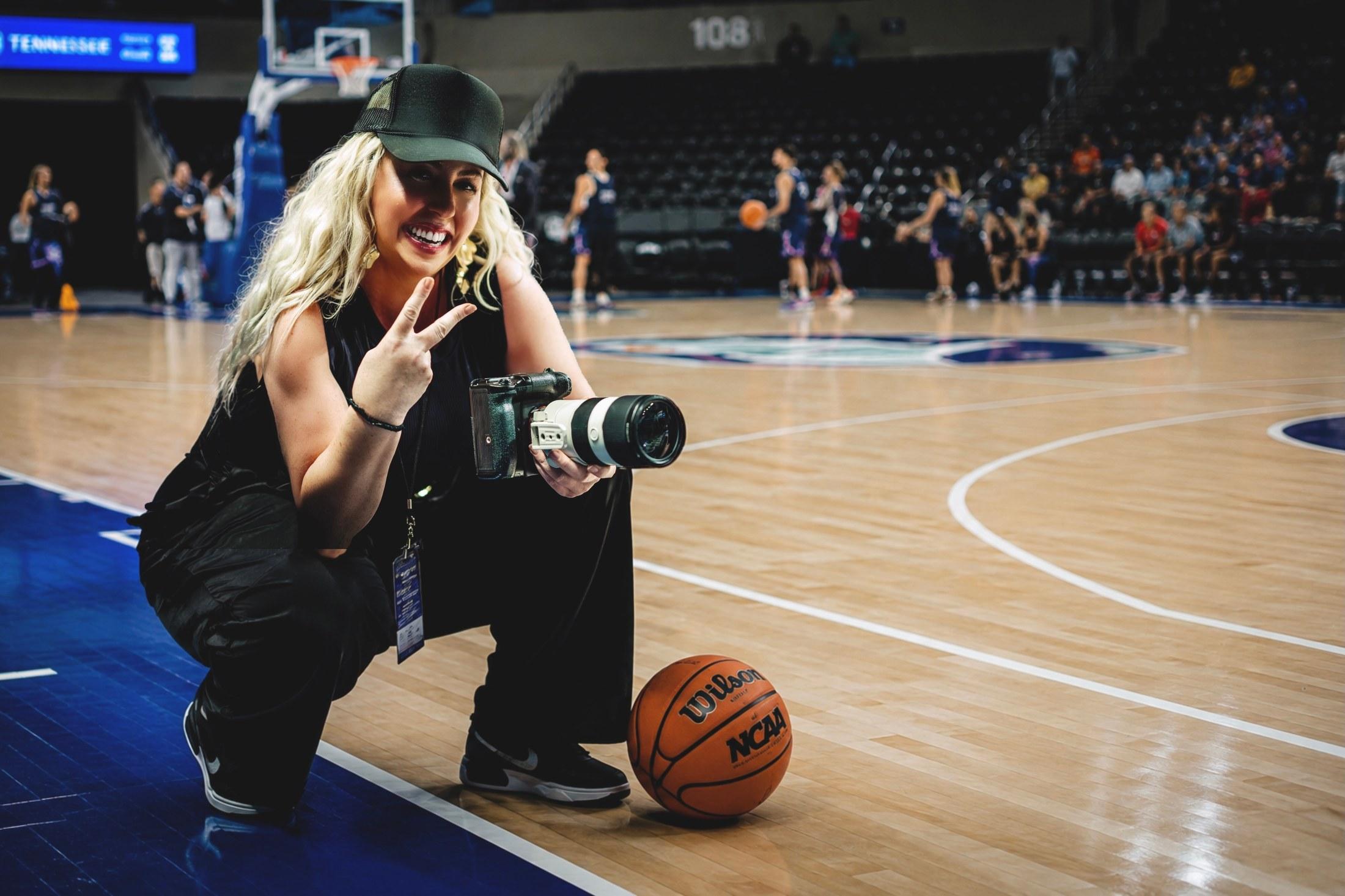 Heather kneeling courtside with her camera and a basketball, smiling at the camera during a game.