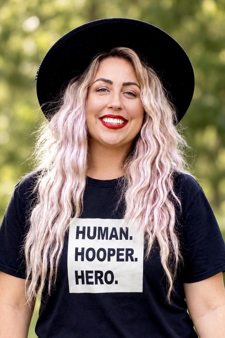 Heather Cleaves smiling outdoors in a black wide-brim hat and a black T-shirt that reads HUMAN. HOOPER. HERO.