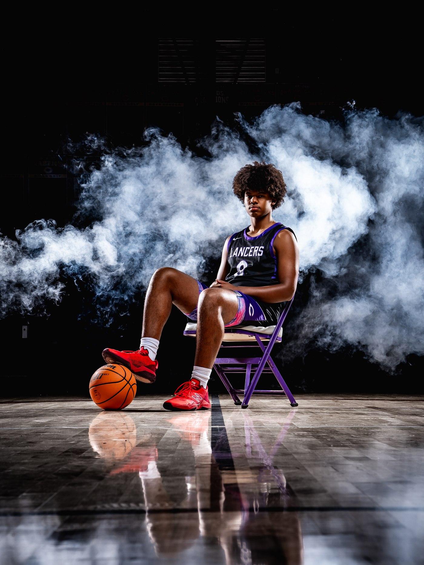 A Lancers basketball player in a purple and black jersey seated on a folding chair, smoke swirling behind him.