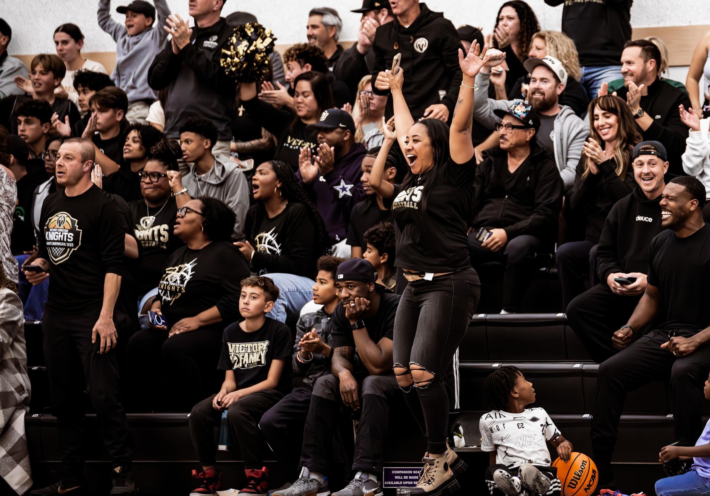 A packed gym of parents and family on their feet cheering, arms raised.